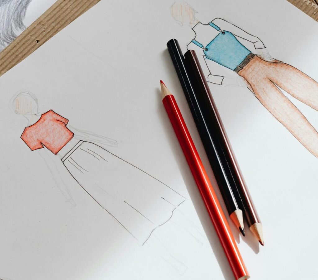 Artistic fashion design sketches on paper accompanied by colored pencils on a wooden surface.