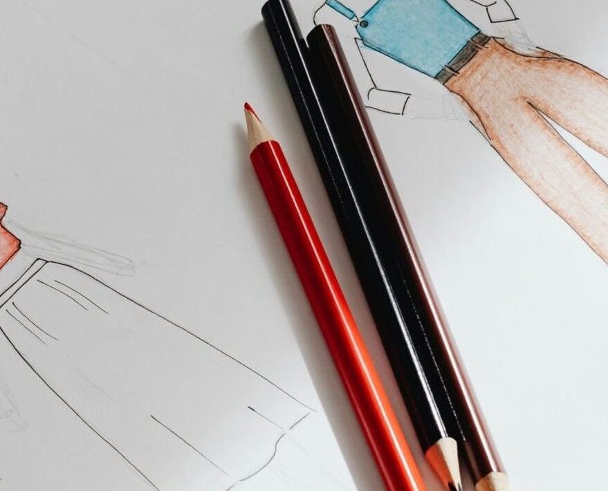 Artistic fashion design sketches on paper accompanied by colored pencils on a wooden surface.